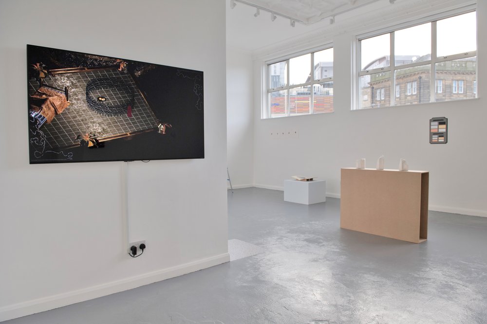 A view of a white cube gallery with grey floors. Part of a wall in the foreground has a large tv monitor on it with a scene from a video game, a dark digital image. In the middle of the space is a cream coloured plinth with three white plaster objects on it. Behind that a small plaque on the wall and a smaller plinth with a book on top. the space has two large windows letting in light.