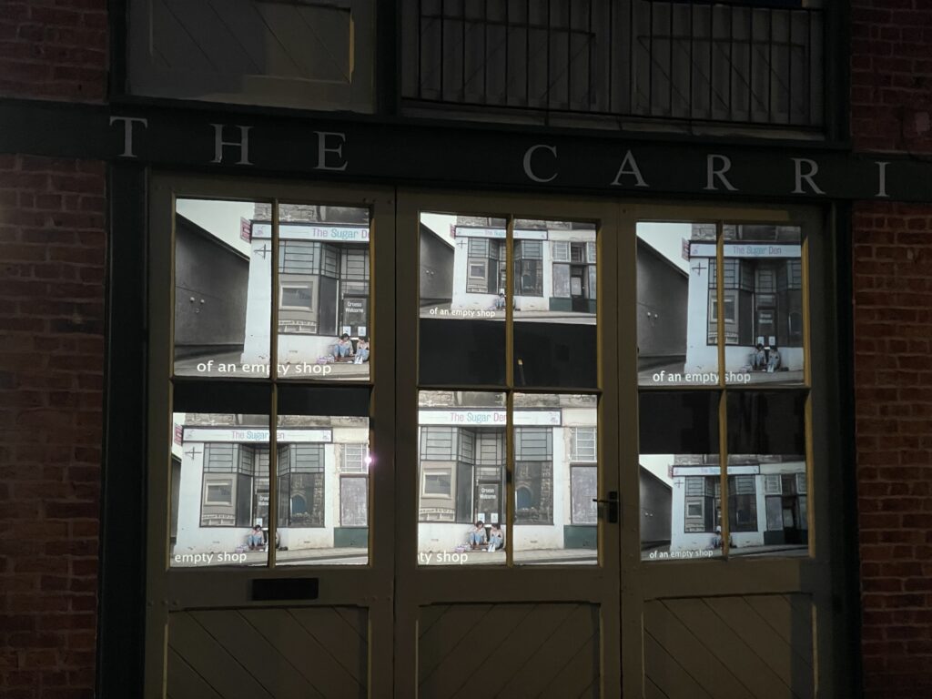 Film depicting a shop front with two women sitting in its doorway is viewed through glass panelled doors.