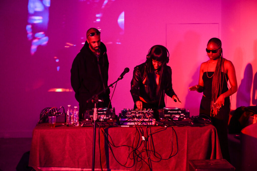 Three people stand behind decks and mixing desk in an industrial space lit red