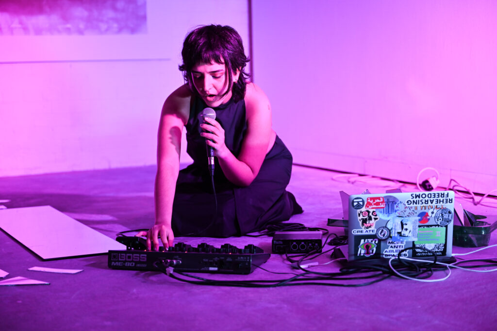A person kneels, microphone in hand, manipulating audio equipment in an industrial space lit fuschia