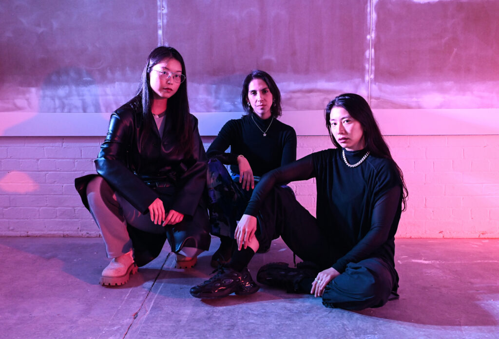 Three people in black clothing pose in an industrial space lit fuschia