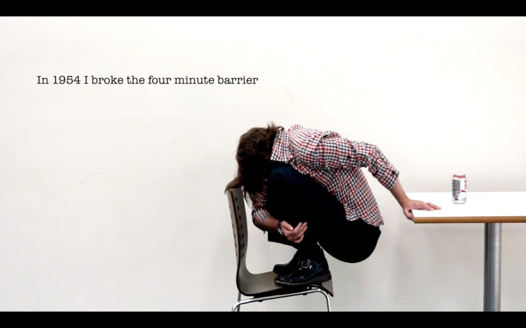 A person curled up back to front on a canteen chair, leaning their hand back against a canteen table where a silver drinks can is visible. Text on screen reads 'In 1954 I broke the four minute barrier'