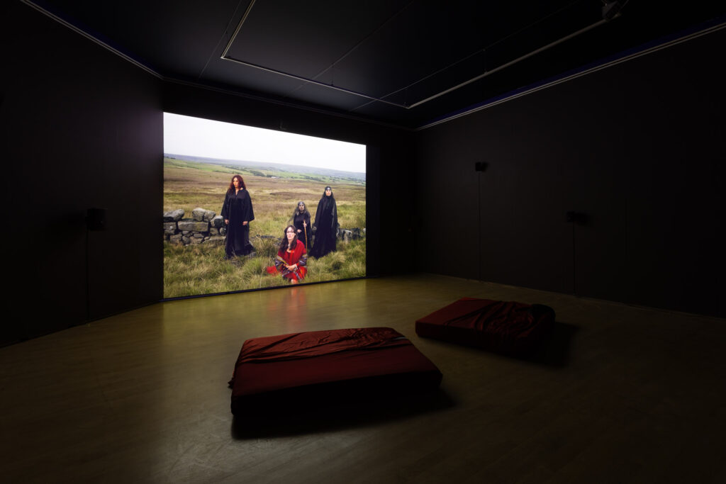The film screen shows the deep greens and browns of north English moorland where three women in dark robes stand around a seated woman in a red robe