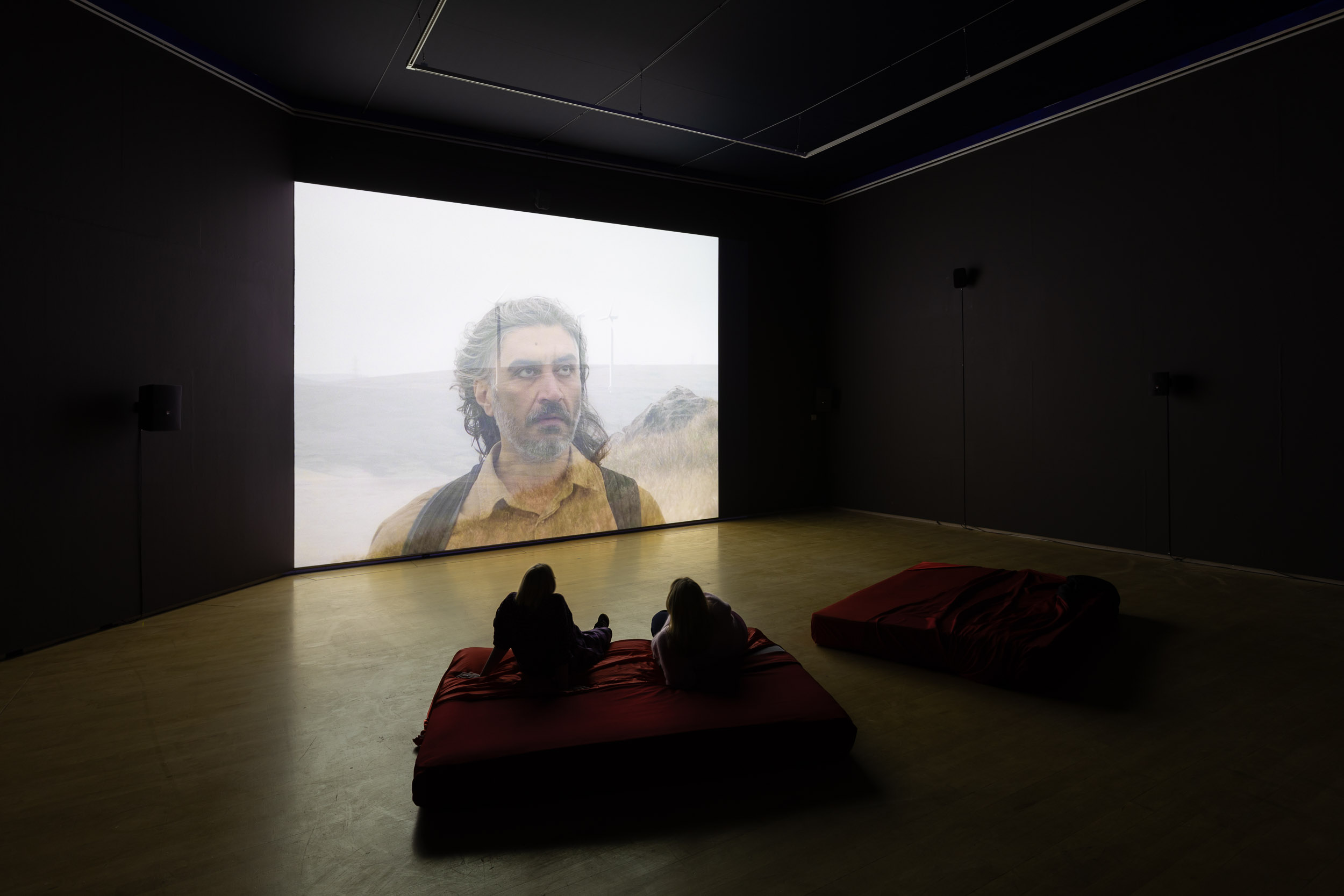 A dark room with deep red bean bags in front of a large film screen which shows a man with white in his beard, semi-transparent laid over the image of a hill