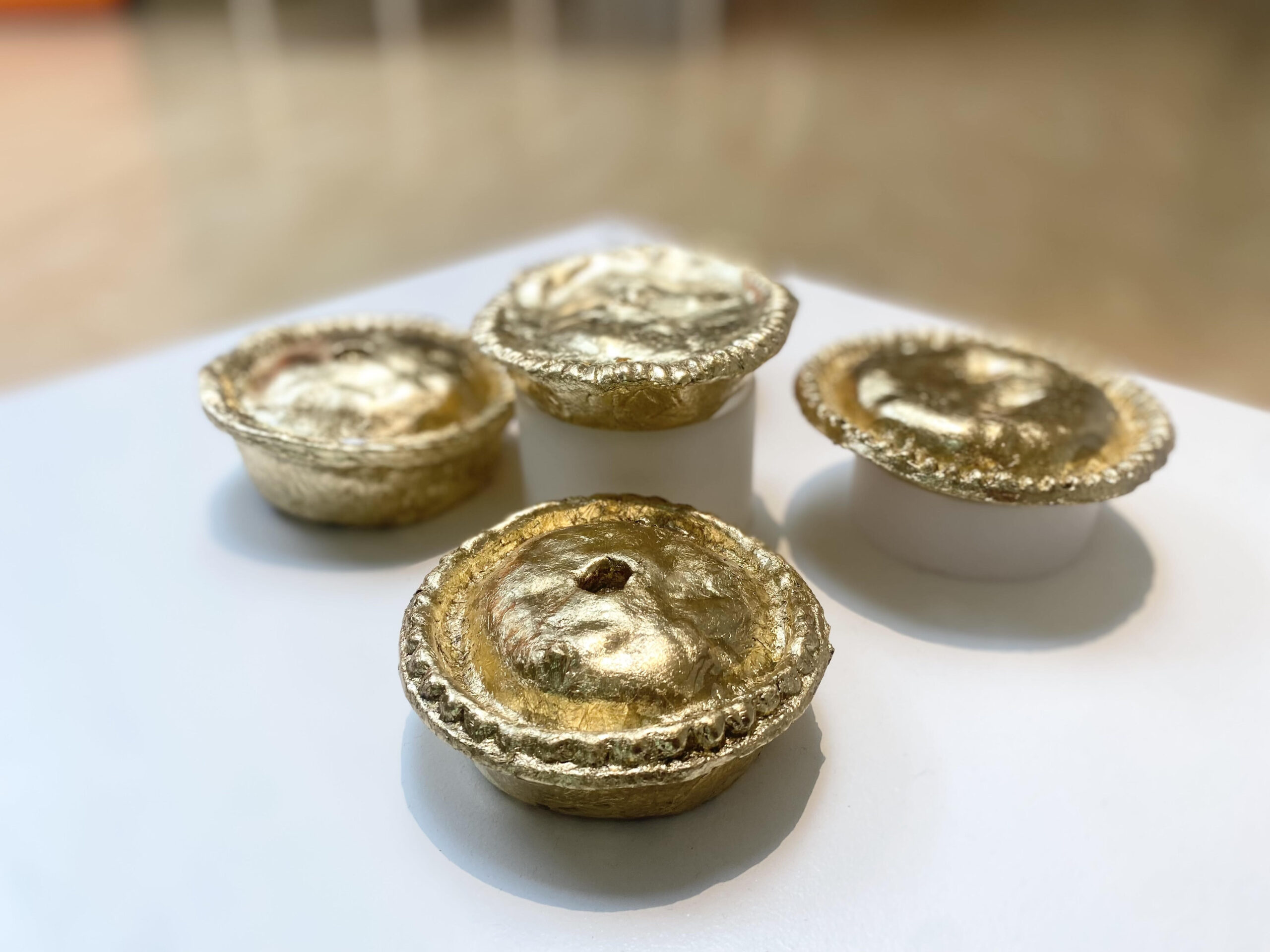 A close-up photograph of four pies which have been covered in gold leaf positioned on a white plinth.
