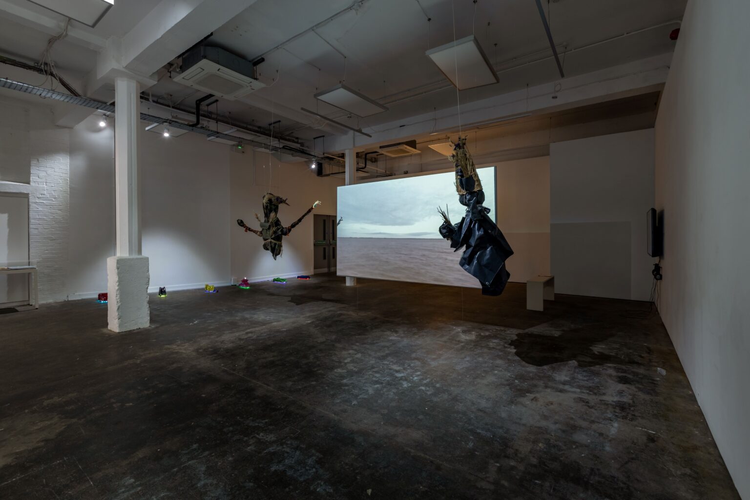 A gallery space with a hanging screen in the centre showing footage of a broad river flowing past. Flanking the screens are two large puppet-like forms also suspended from the ceiling. Along the wall in the background are lined brightly-coloured model bridges, under-lit with coloured LEDs.