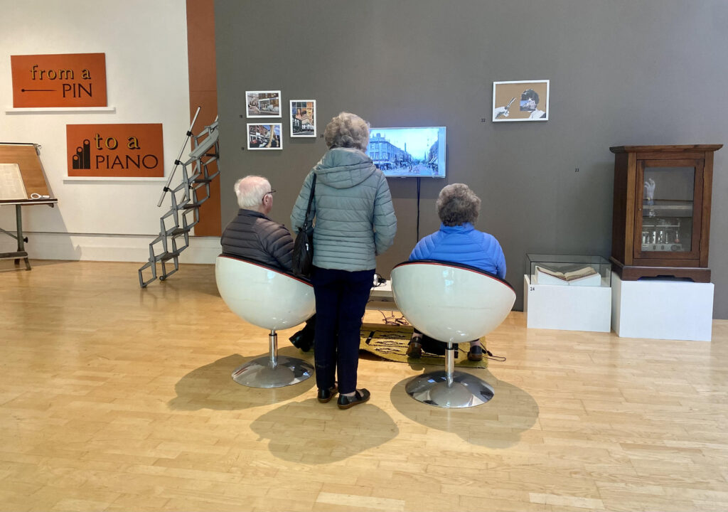 Three visitors at an exhibition watch a screen with their backs to the camera, two are seated in 1960s style egg chairs whilst another stands between them. They appear to be older white people. There are other works in a variety of media on the surrounding walls.