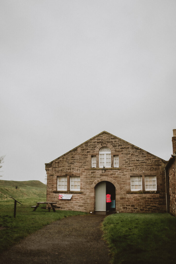 A stone building in a rural setting. It has an arched doorway with a curved path leading up to it. there is a picnic bench outside.