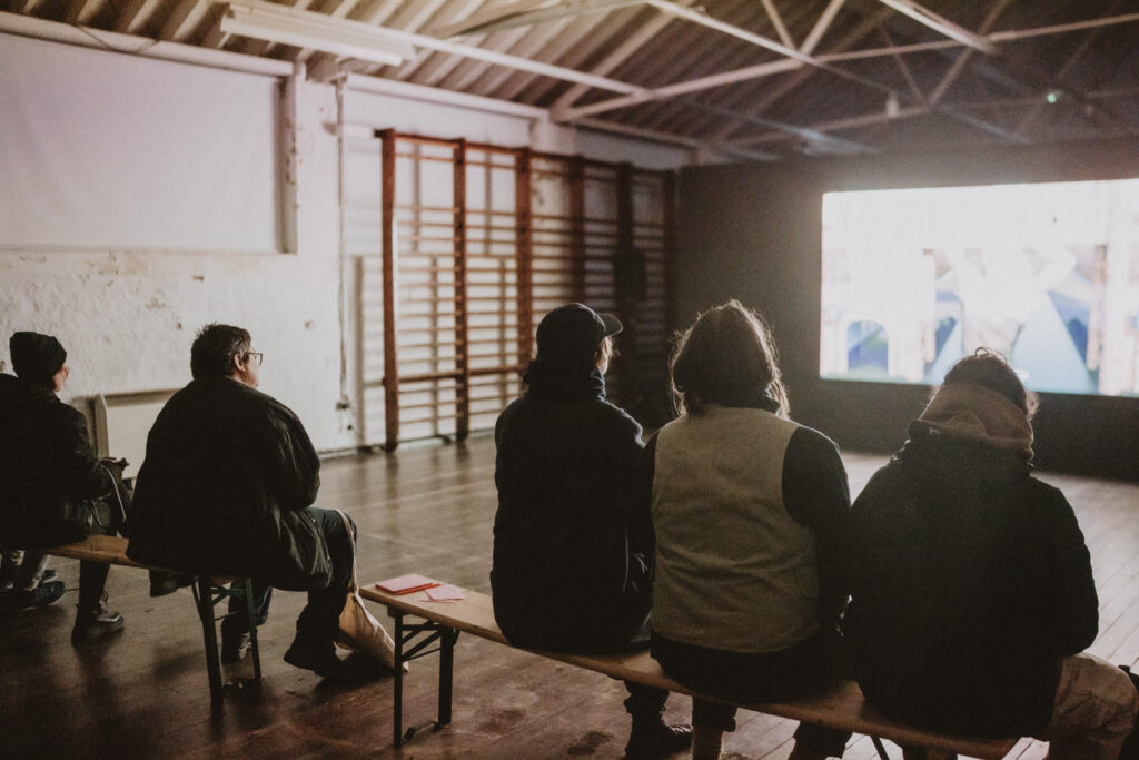A group of five people sat on benches in a former gymnasium. They are watching a film projected on the wall. The image is very bright and difficult to see what they are watching.