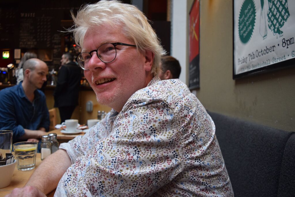A white man with white hair and stubble sits smiling, looking ast the camera. He is wearing black rimmed glasses and a light coloured floral shirt. He is in a bar or restaurant.