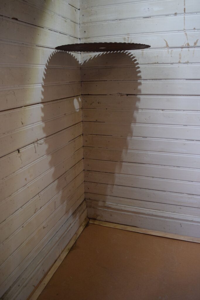 A photo of a saw mill blade wedged in the corner of two walls and casting a shadow in the shape of a jagged love heart.