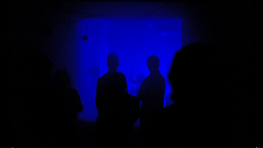 A dark room lit blue with a standing audience silhouetted and a drummer visible at the back wall
