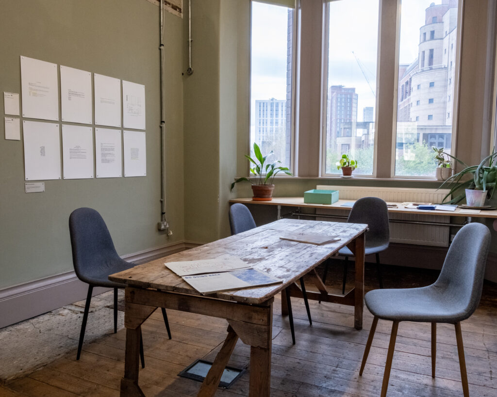 A table with four chairs arranged around it in a room with a window. On the wall to the left are approximately ten sheets of A3 sized paper with what looks like text, pinned in a grid. there are three booklets on the table. There is another table behind, by the window, with more printed material and some potted houseplants.