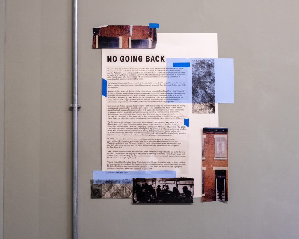 A collage made directly on the grey gallery wall, very close to a stainless steel pipe. The collage is around A3 in total size and is made up of an A4 sheet of text with No Going Back, in large font at the top. There are five fragments of photographs and two pieces of translucent blue paper, taped onto the larger sheet, aound the outside mainly, with small pieces of blue tape.