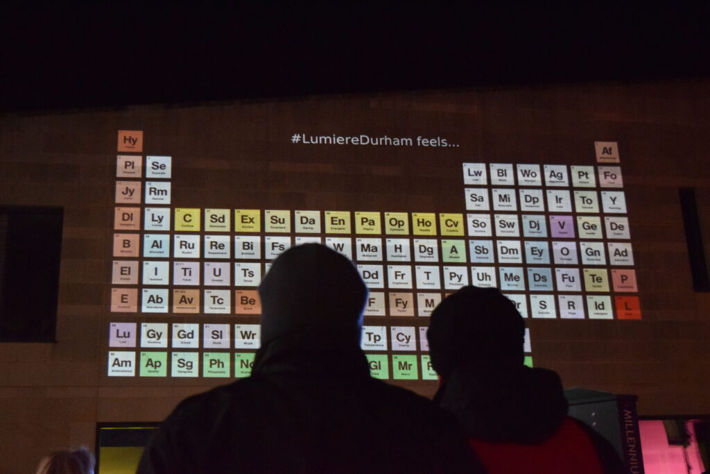 We see a couple, from behind looking up at a projection on a high wall. The image projected looks like the periodic table of elements but on closer inspection you see each element is an emotion.