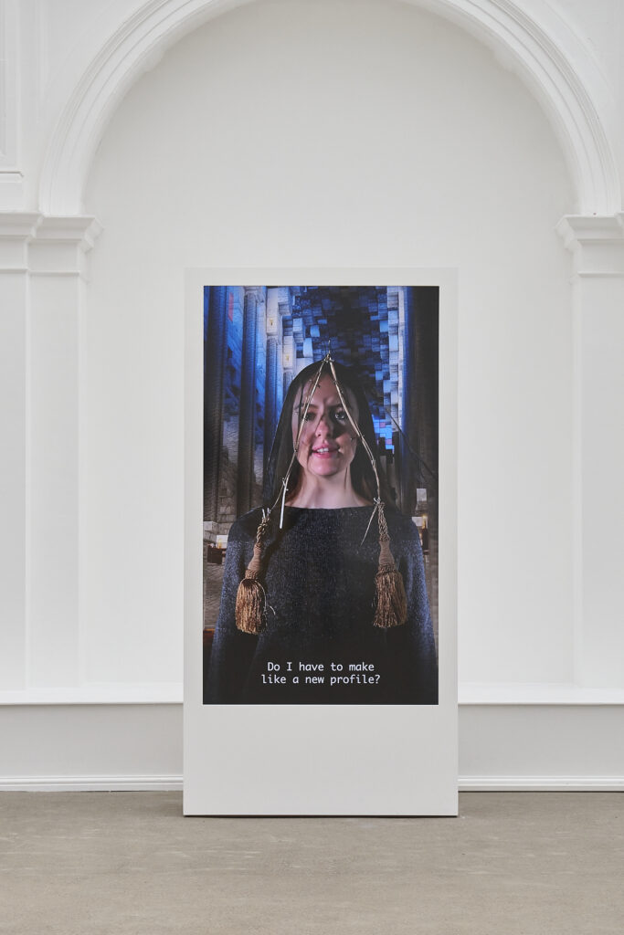 A large portrait-orientated screen in a large white space, framed by an arched recess in the wall behind. The screen shows a person in a head covering in a digital, church-like space. A caption reads 'Do I have to make, like, a new profile?'