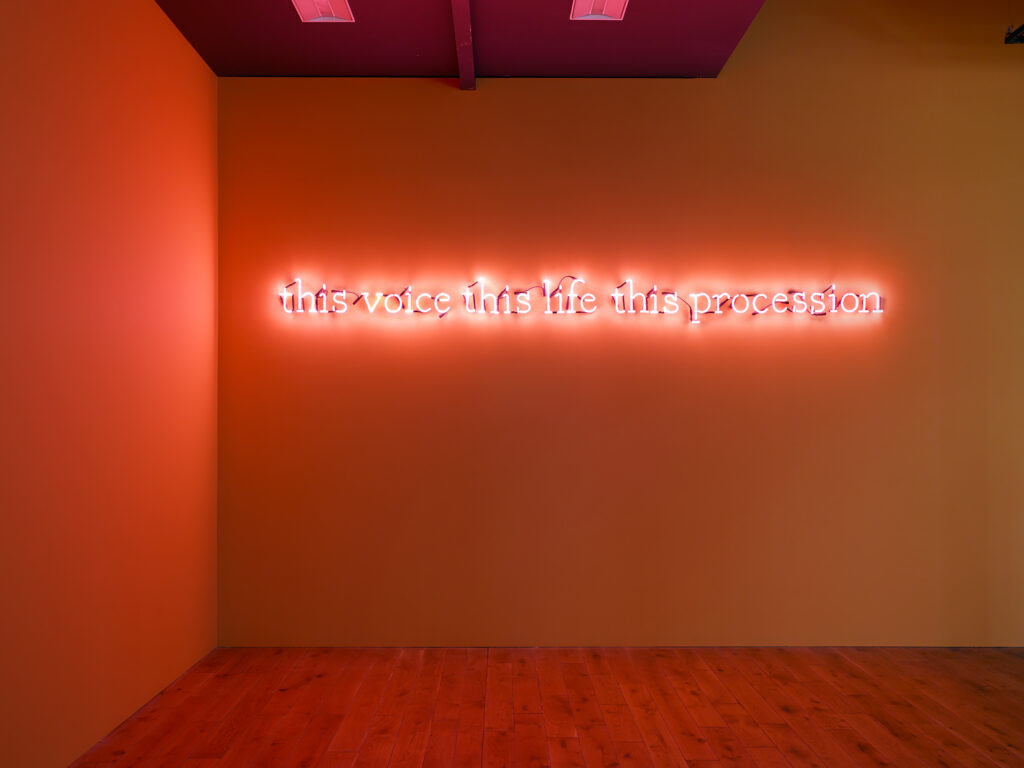 A room with wooden floors and mustard walls with a neon text light illuminated in pink which reads: this voice this life this procession