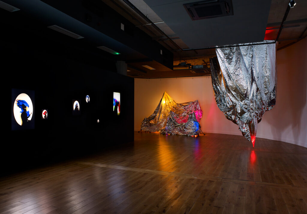 In a darkened gallery space the side wall to the left is black with illuminated silhouettes displayed, and straight ahead are two huge silver foil structures lit up multicoloured at various points
