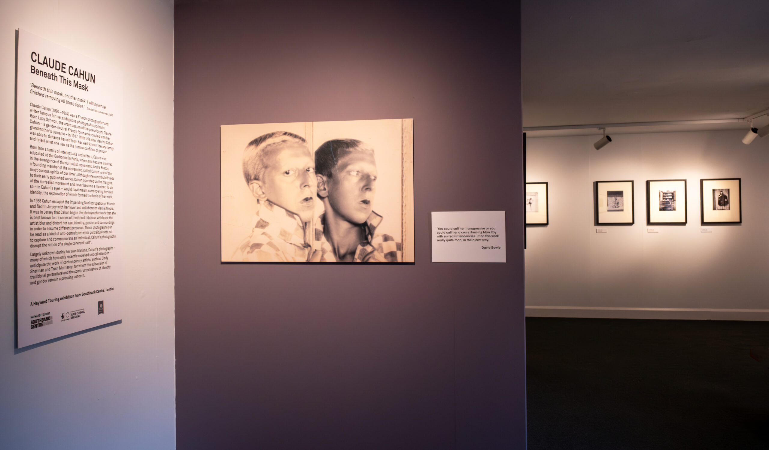 A gallery view with introductory wall text on the left and straight ahead one of Cahun's mirror portraits displayed on a deep purple wall