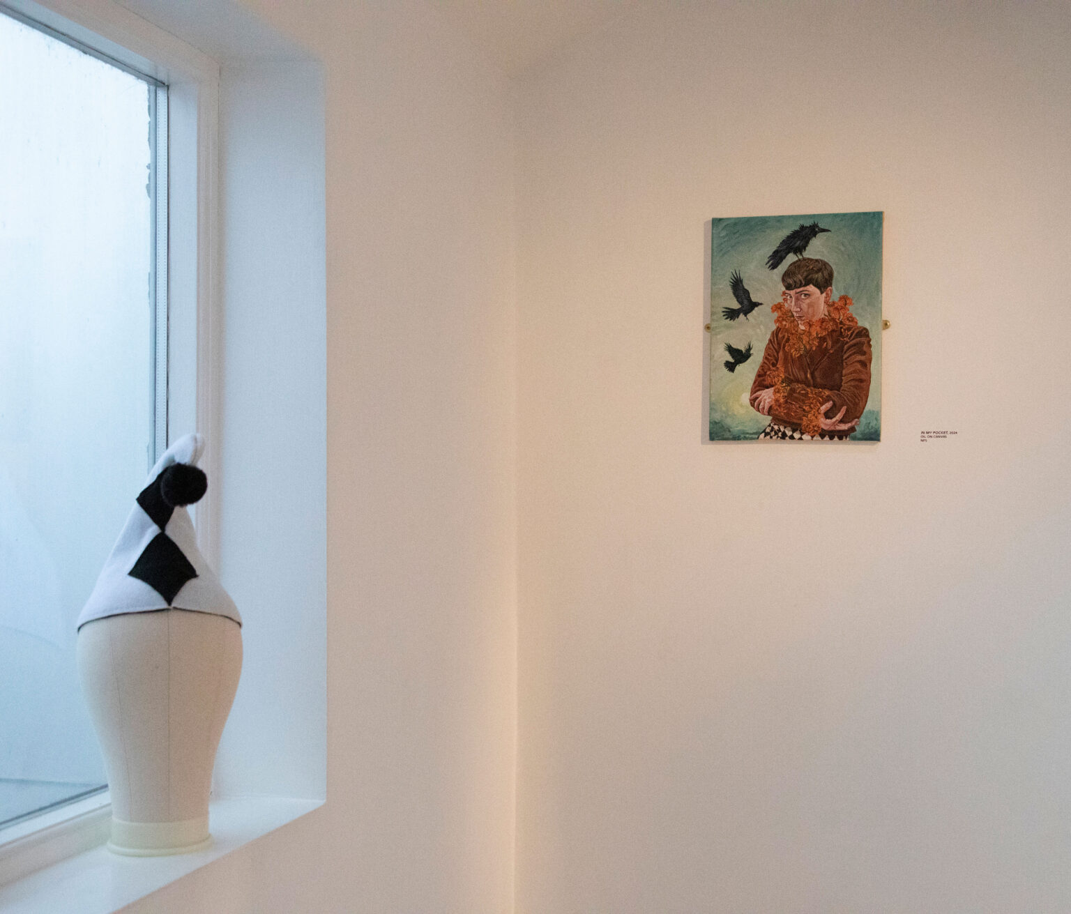 A white room with a window to the left, on the window ledge a model head wears a pierrot clown's hat and on the wall ahead a small painting of Jude in orange clothing with crows flying around her head