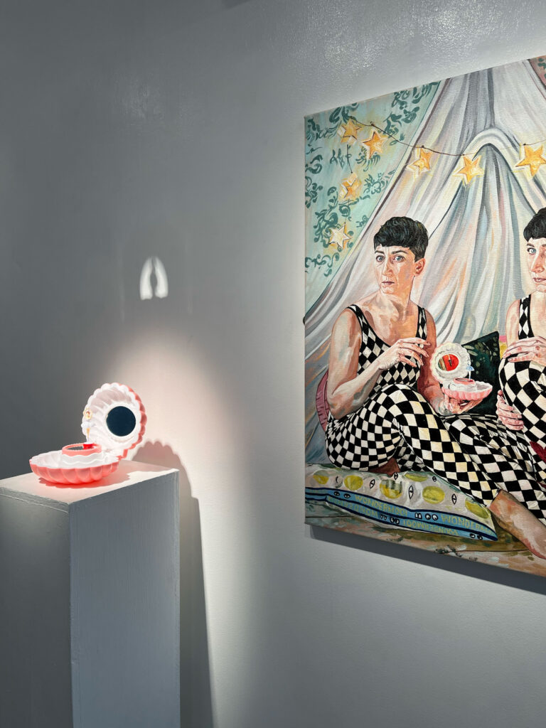 On a plinth is a well-lit clamshell object, opened, with white inside and pink outside, and on the wall behind it a large painting of two Judes in black and white diamond leotards holding the same object