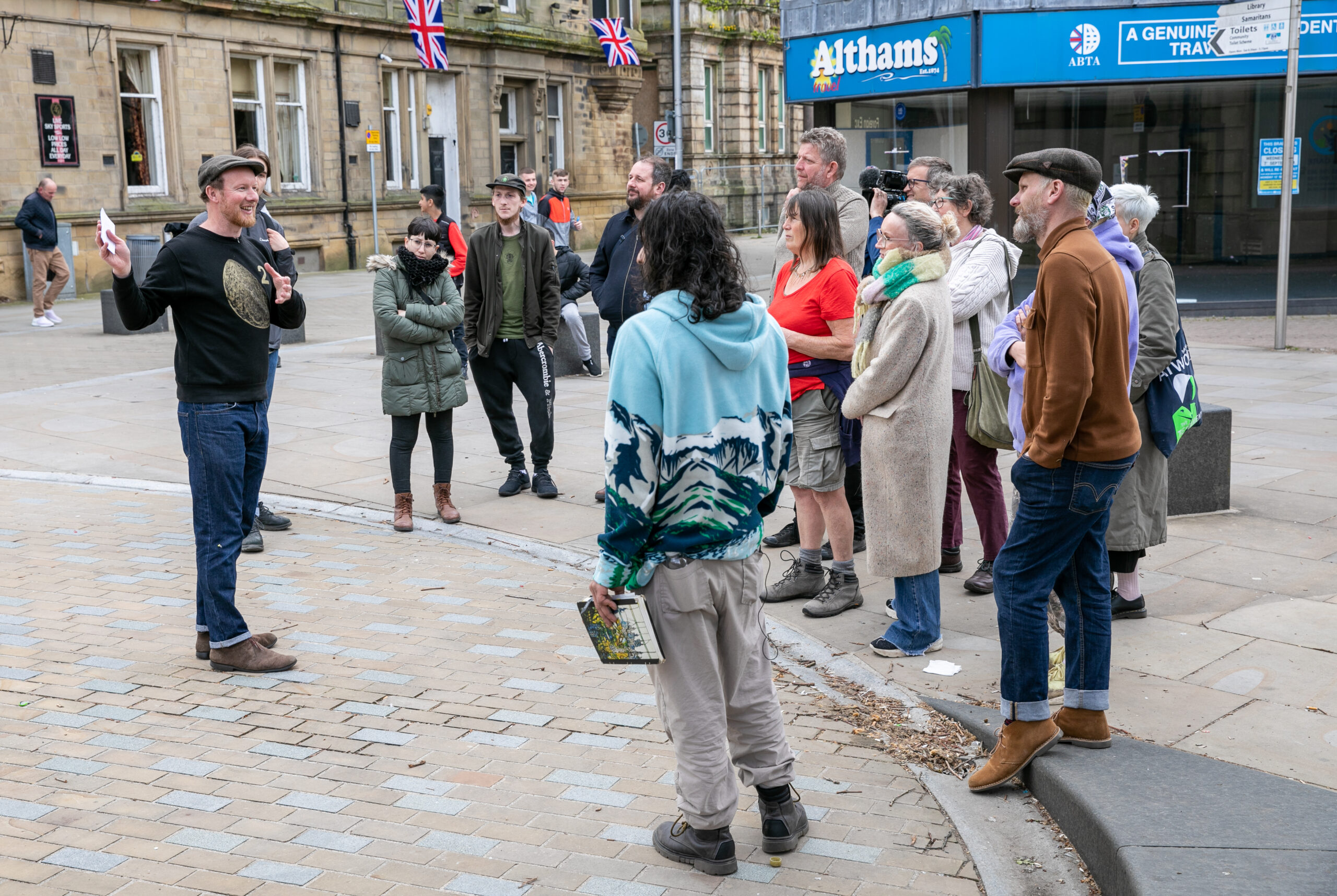 A group of people of varying ages gathered in a public square or street in front of an old shop front