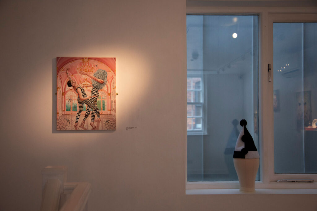 A white wall with a small painting to the left that shows two judes in black and white leaotards dancing together, the one on the right has a blue wig that covers her entire head and the top of her torso. To the right a clown's black and white hat on display in the window.