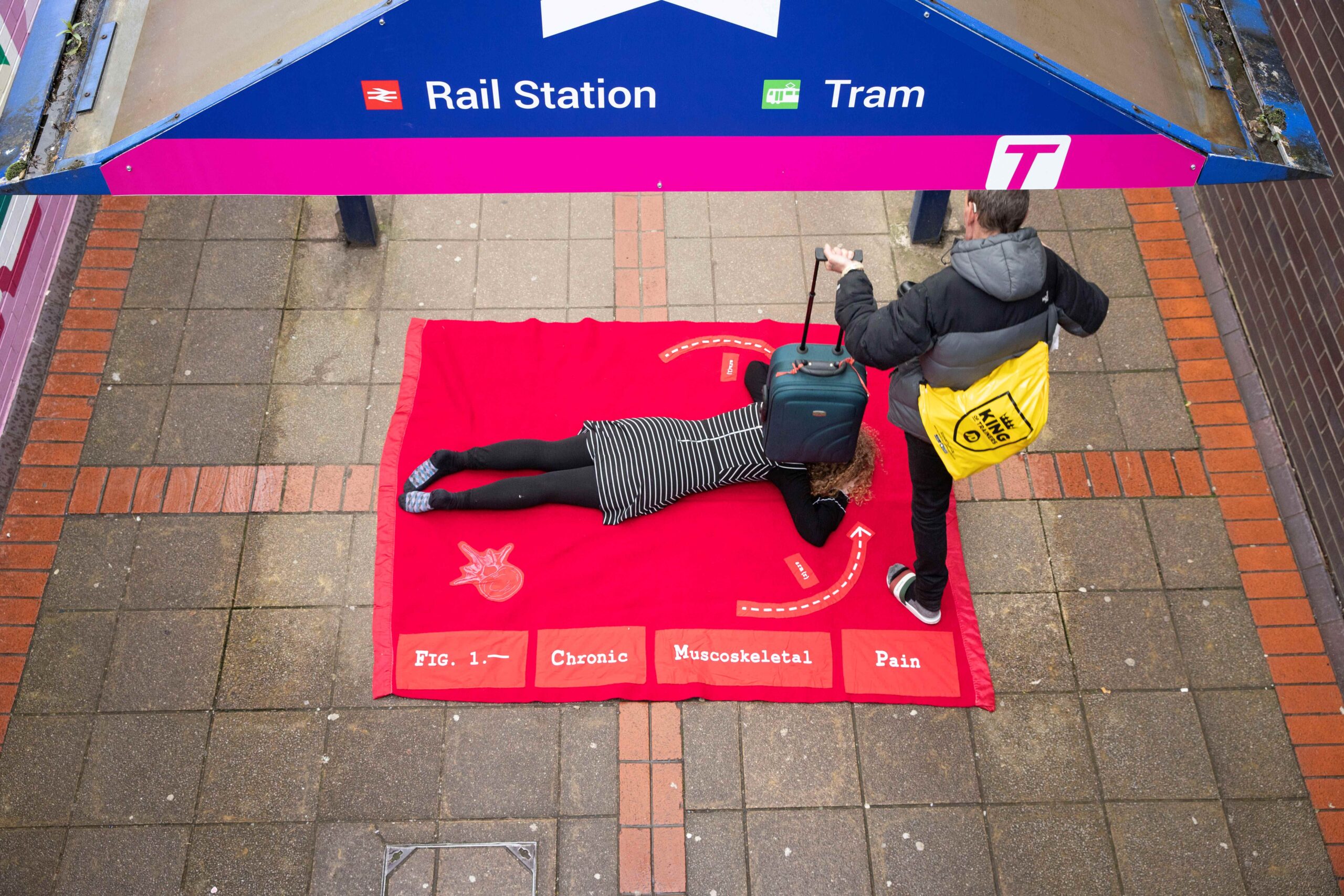 A woman lies on a soft-looking, red blanket on the floor in the middle of a railway concourse; a commuter carrying a rolling suitcase steps over her.
