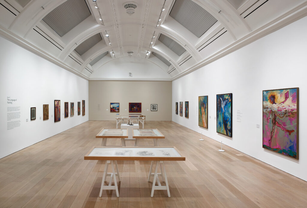 A long view of the gallery space with paintings on the left, back and right walls, and in the centre of the space low tables with drawings