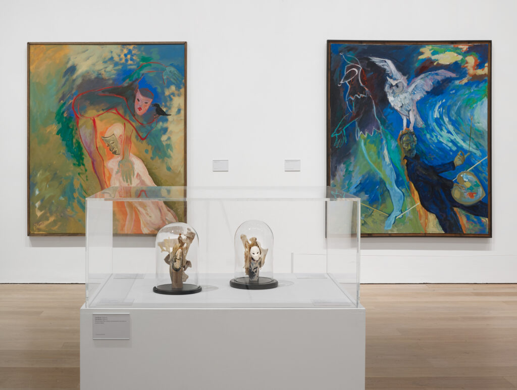 Two paintings on a white wall, both with blues and greens, showing figures in abstract space, on the right an image of the painter with palette and brush in hand being carried away by an owl, and in the foreground two small assemblage sculptures in a vitrine