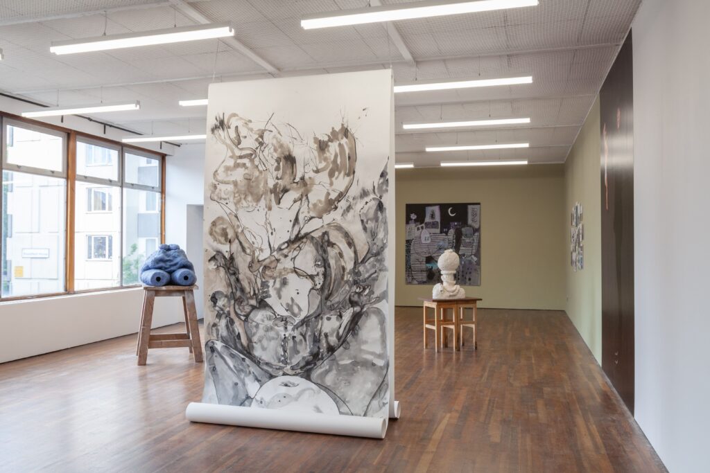 A brightly lit gallery space. In the middle foreground is a large painting on paper, suspended from ceiling to floor. The painting is a watery figure. Behind this on either side are sculptures on small wooden tables. there is a large painting on the far wall.