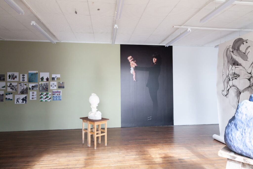 There is a large photo pasted onto the gallery wall, from floor to ceiling. It shows a woman with medium length brown hair and wearing a suit, holding a baby at arms length.