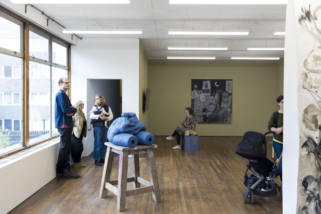 A gallery space with a large bright window. There are five people looking at art works and talking. In the foreground is a blue sculpture on a wooden plinth structure. On the wall in the background is a large painting of a landscape at night.