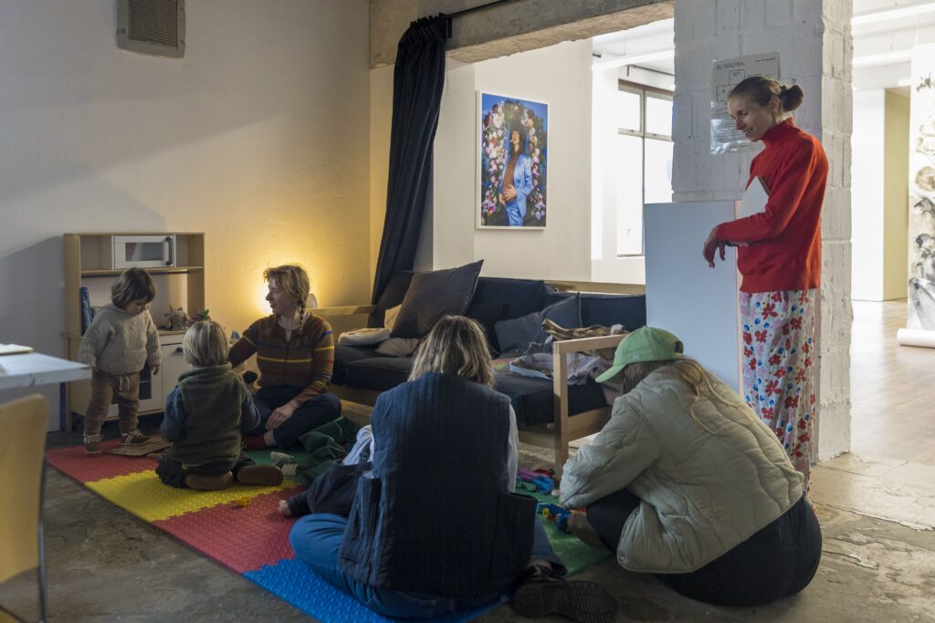 Two children and three adults are playing on a mat of coloured tiles. There is a sofa behind them, and on the wall behind that is a photograph of a woman smoking a cigarette with her hand on a pregnent belly.