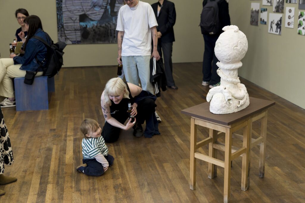 A baby and an adult are smiling as they look at a white plaster sculpture on a small wooden table. There are other people in the gallery space chatting and looking at art.