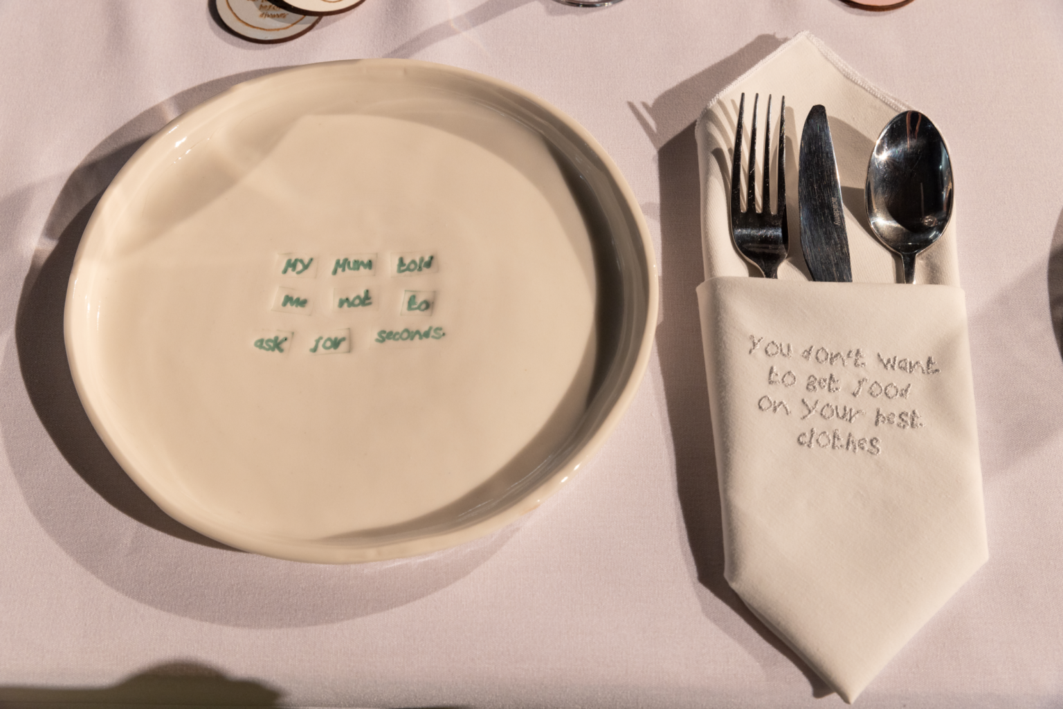 a cream coloured glazed plate is stamped with the words 'my mum told me not to ask for seconds' in a pale sage green, to the right hand side an embroidered napkin is folded around a set of cutlery, the embroidered word say 'you don't want to get food on your best clothes', they are all laid on a white tablecloth and lit from the side which creates shadows