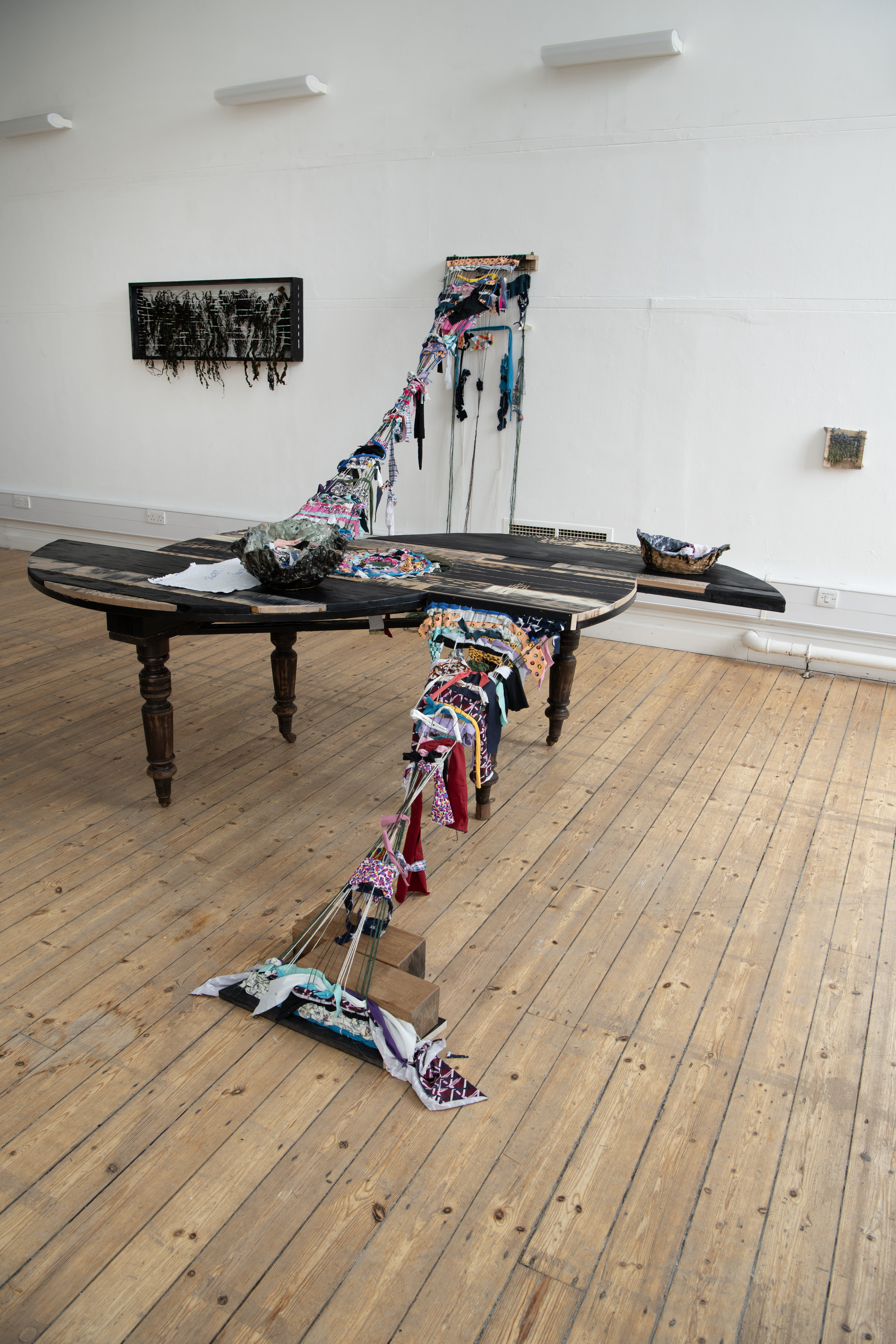 A large sculpture made from a cut up round dining table and other found materials such as clothing and household objects. It is attached to the wall and the floor.