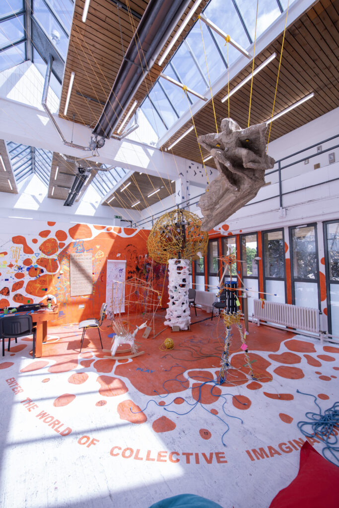 A huge messy installation of found objects assembled into a network. The floor and walls partiall painted in orange blobs.