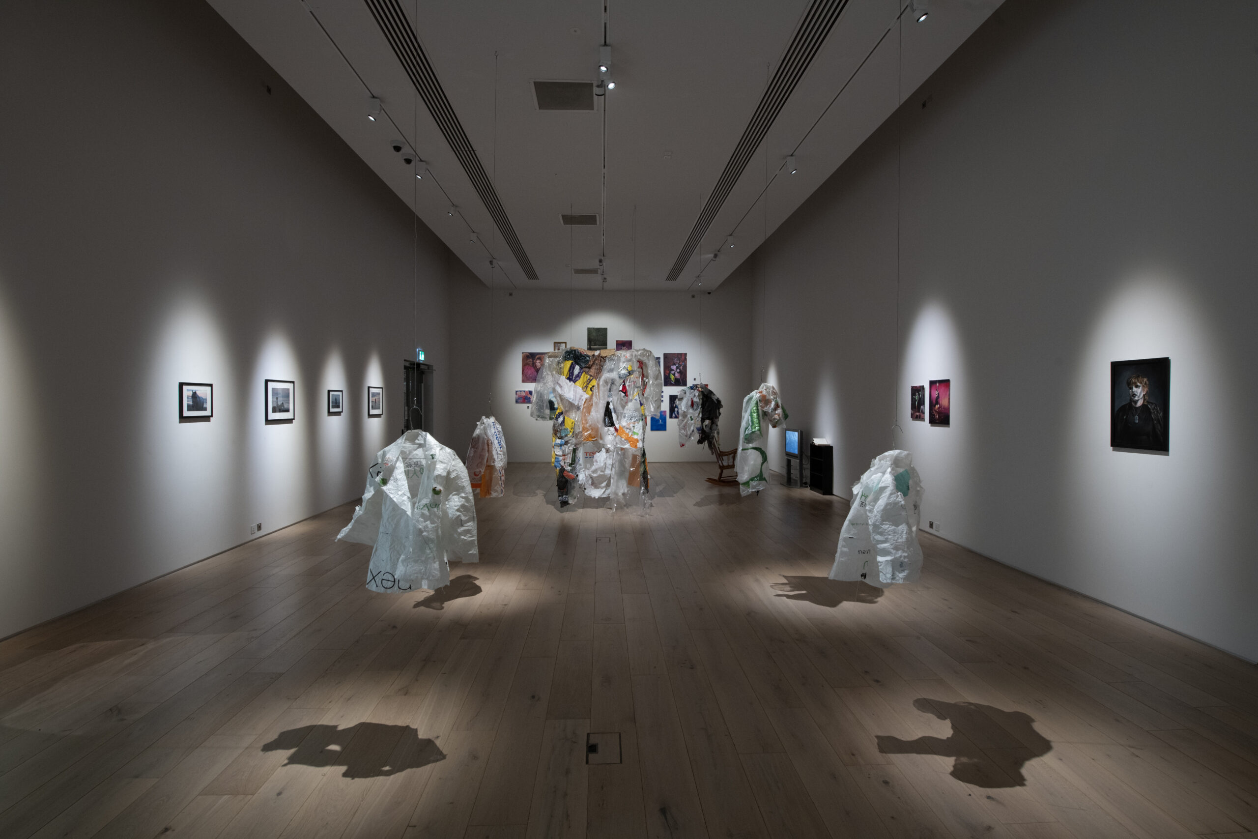 A large gallery space with several garments made out of waste materials like plastic bags and packaging suspended from the ceiling. Each garment is spotlit in the dim space.