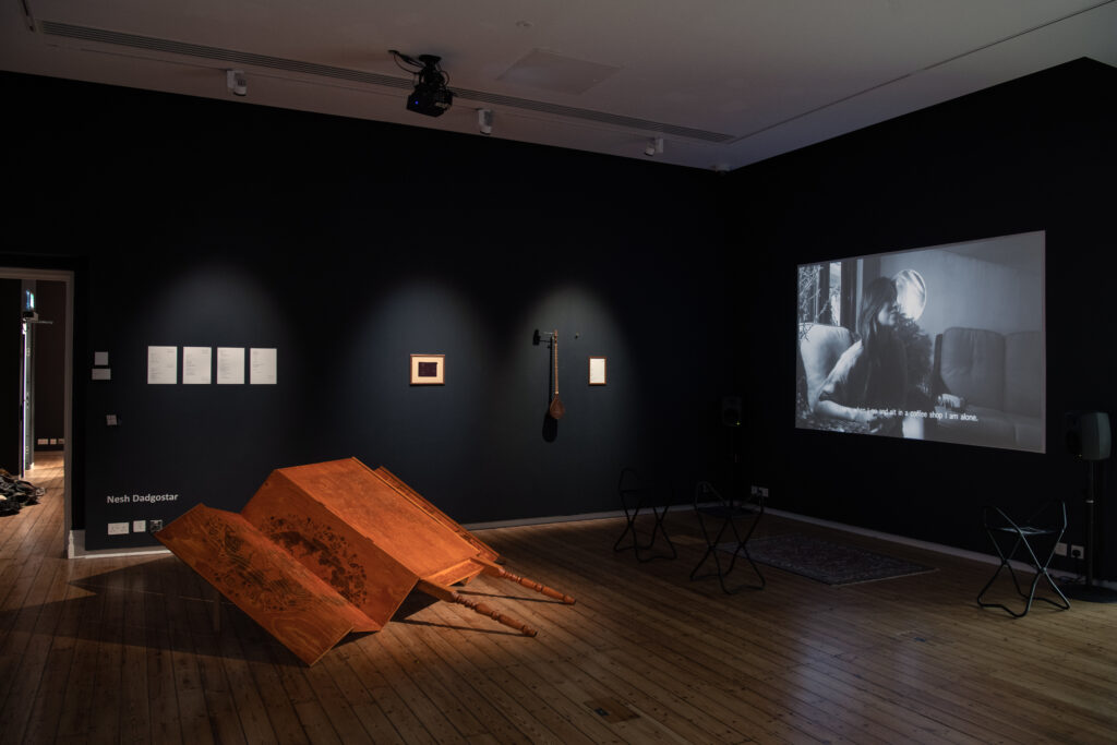A gallery space with wooden floors and walls painted black. Small works are spotlit on the wall. On the adjacent wall a projected image. On the floor in front a large sculptural form, like an orange screen tipped over.