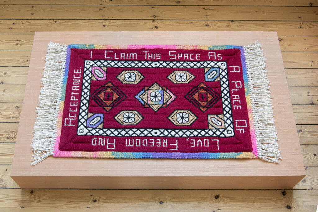 A woven mat with the words 'I claim this space as a place of love, freedom and acceptance' woven in white on a red background. Displayed on a block on the floor.
