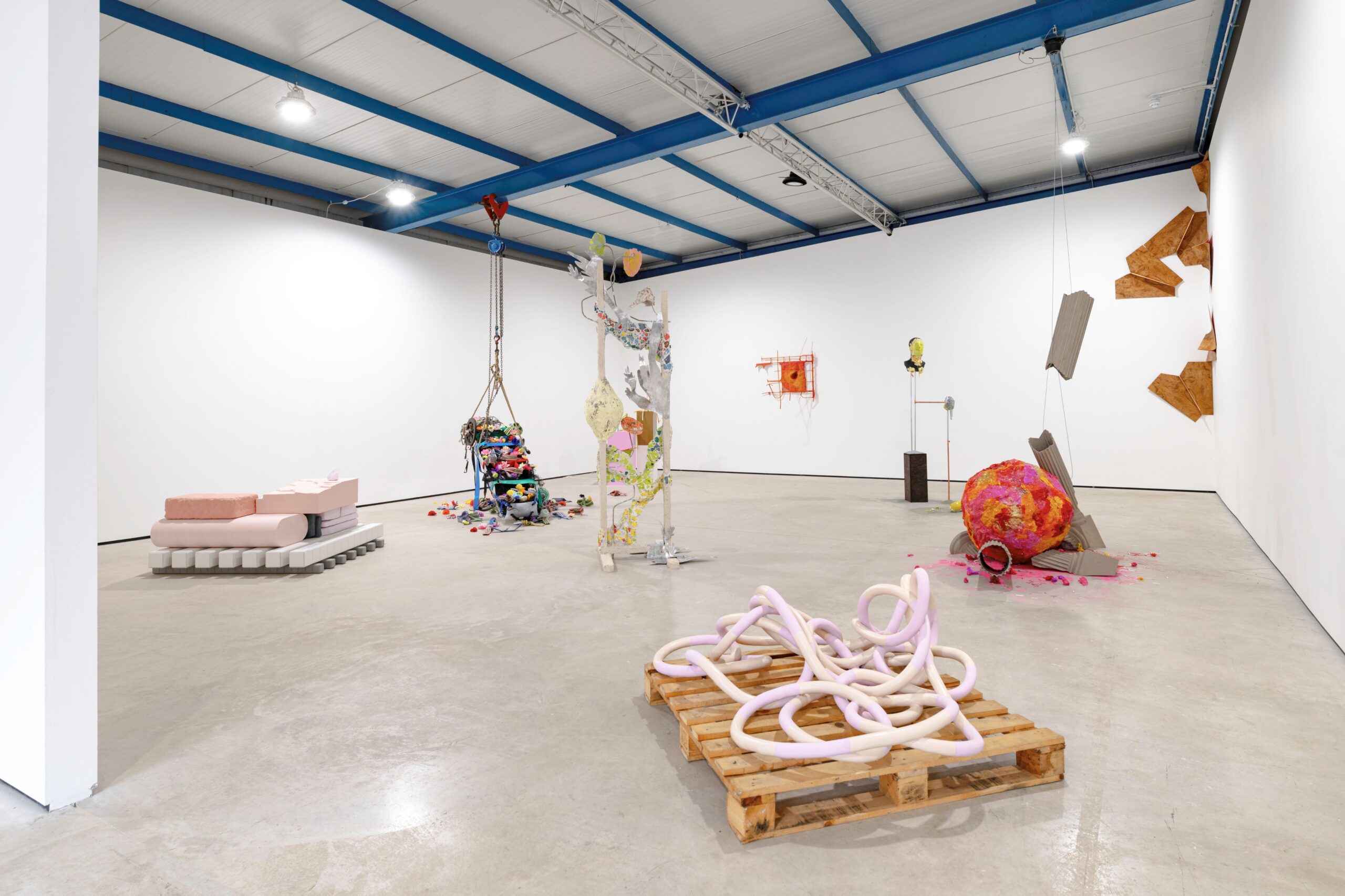 Photograph shows most of the gallery space with works dotted throughout, the works are described in detail in the article but in brief there are a range of shapes and bright colours sat on the floor or tumbling down the walls.