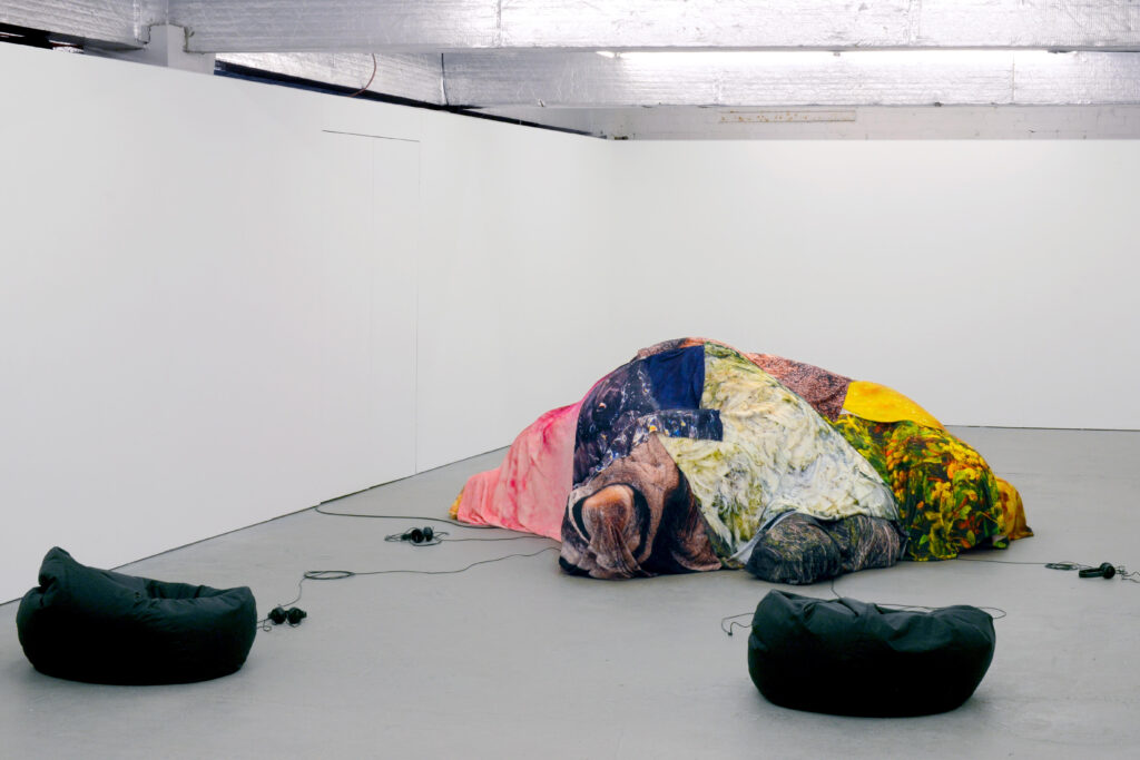 A white walled gallery space with grey floor, in the middle is a mound draped with a patchwork of colourful fabrics, with two black bean bags before it and wired headphones on the floor