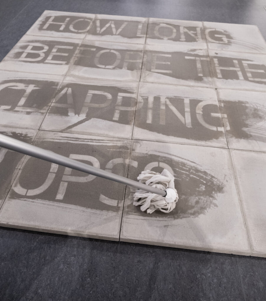 A photograph showing a mop being dragged across concrete tiles revealing the words 'how long before the clapping stops'