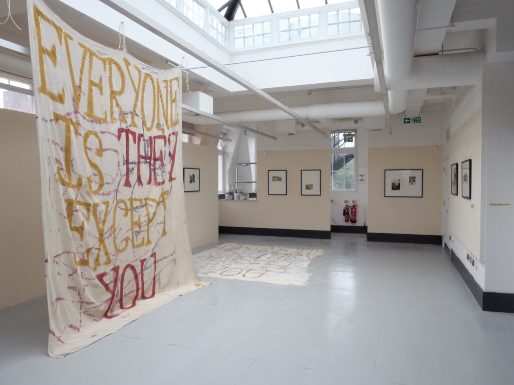 A large sheet hangs from the ceiling with a painted phrase: 'EVERYON IS THEY EXCEPT YOU'. On the back and side walls, painted magnolia, are framed black and white prints.