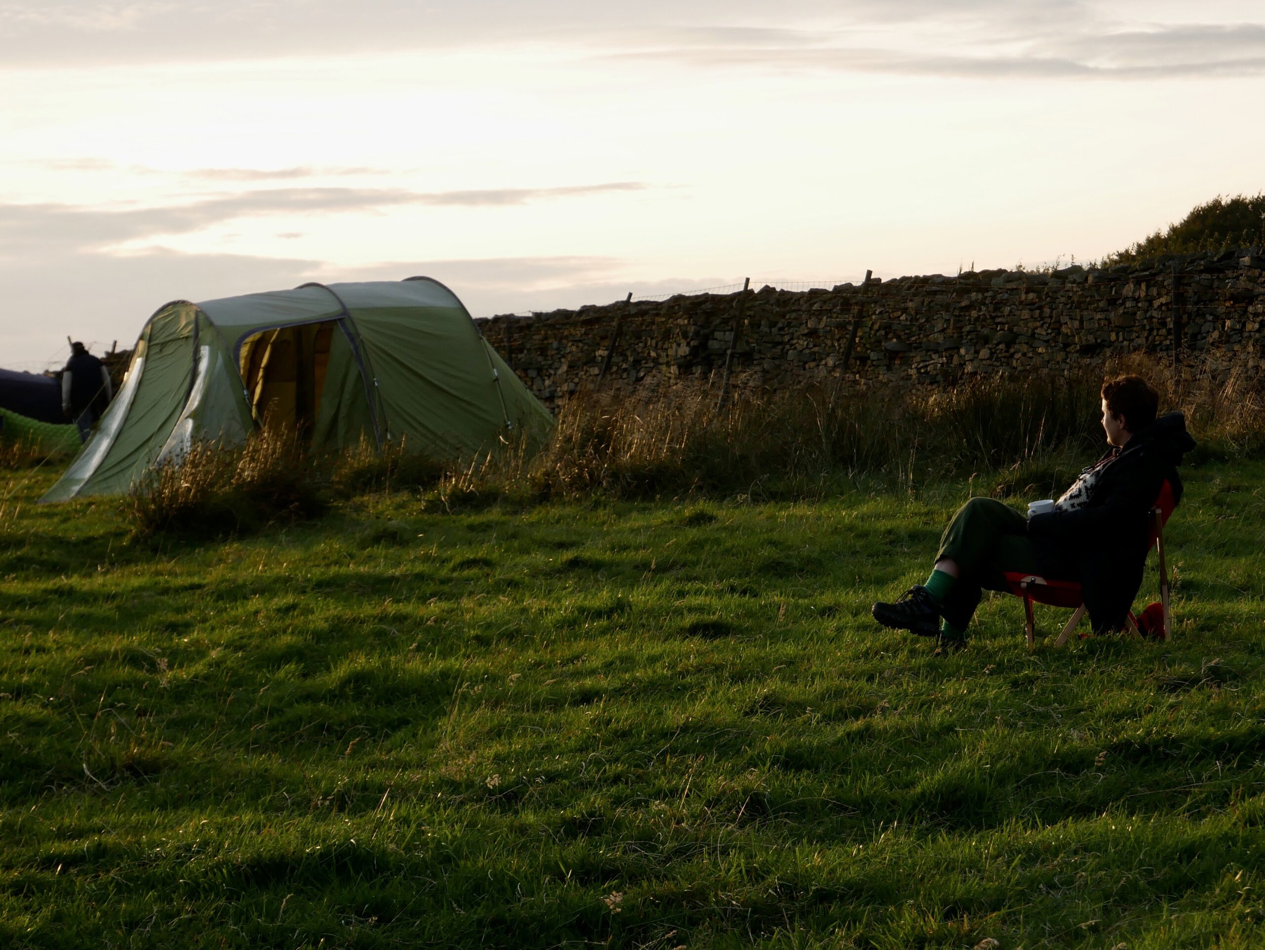 A person sitting in a camping chair in a field with their green tent in the distance to the left at dusk