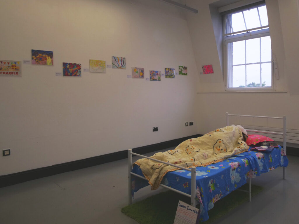 A person asleep in a small bed in the middle of a bright room. Colourful drawings are hung on the back wall.