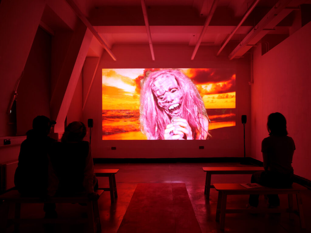 People sit watching a projection screen in a dark room, an orange glow comes from the projected film of a singer in a disturbing mask in front of a sunset beach backdrop