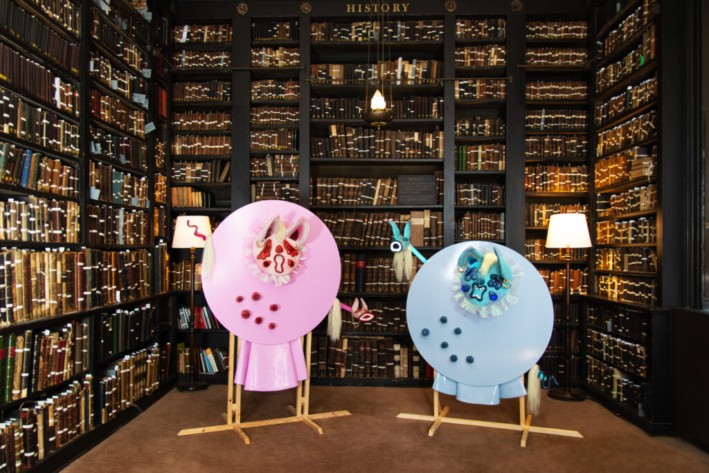 In a dark library room with floor to ceiling wooden shelves covered in leather-bound volumes, two round creatures, on the left baby pink and the right light blue, with sparkling, screaming faces.
