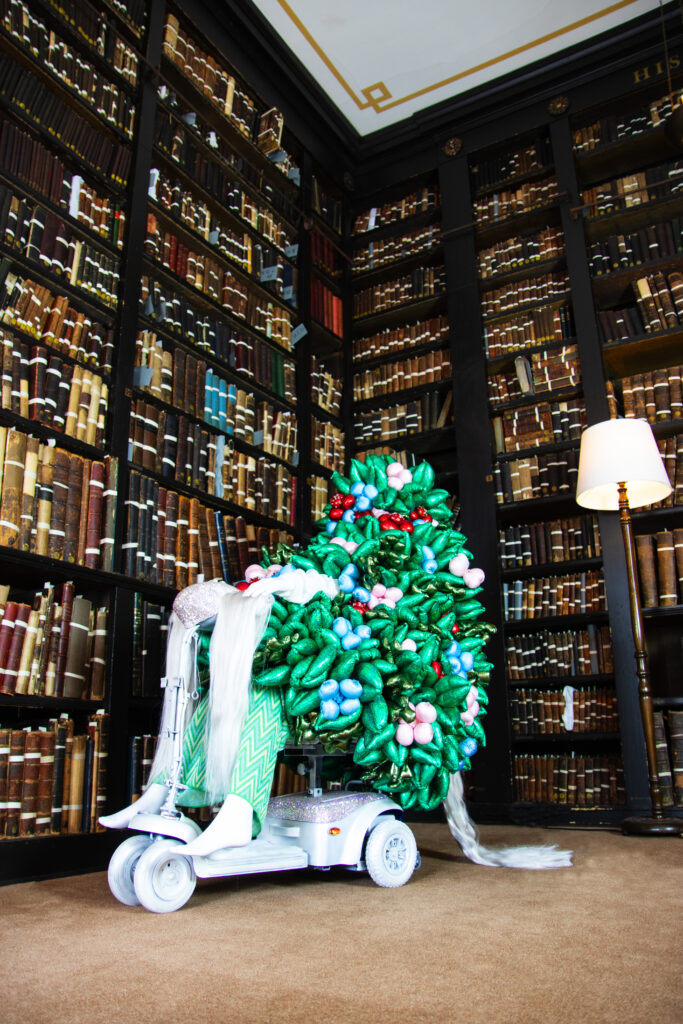 In the dark library room a figure covered head to knees with a plant of round soft looking textile leaves, with blue, red and pink berries nestled in amongst them, sits on a silver mobility scooter, their white feet poking out.